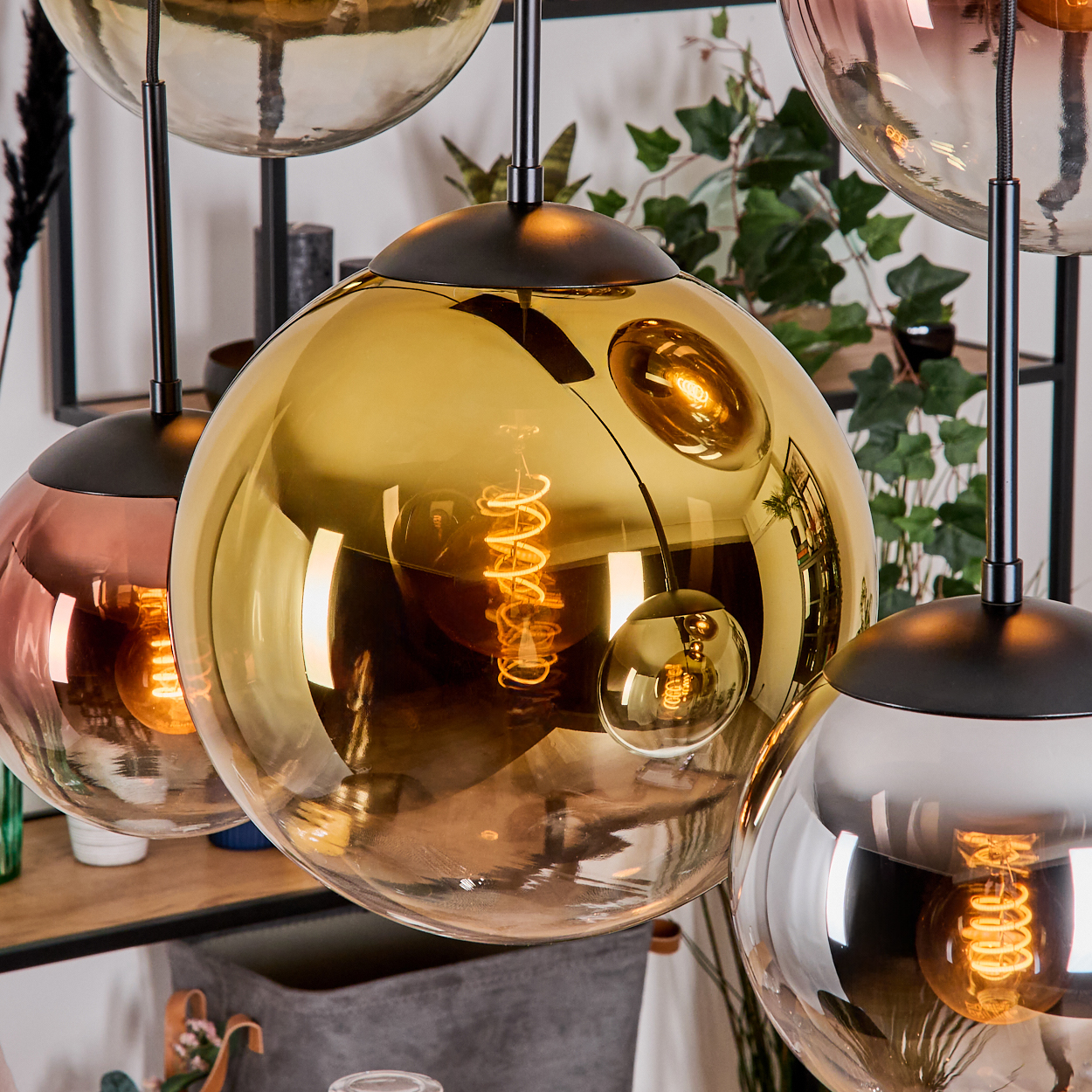 Nahaufnahme mehrerer kugelförmiger Pendelleuchten mit sichtbaren Wendelglühbirnen. Farben reichen von Gold bis Rosa, mit schwarzen Halterungen.
