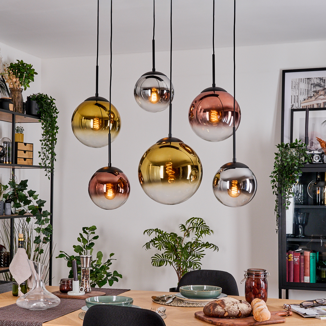 Esszimmer mit hängenden Kugellampen. Tisch mit Essen, Pflanzen und Regalen. Lampen haben Gold-, Silber- und Kupfertöne.