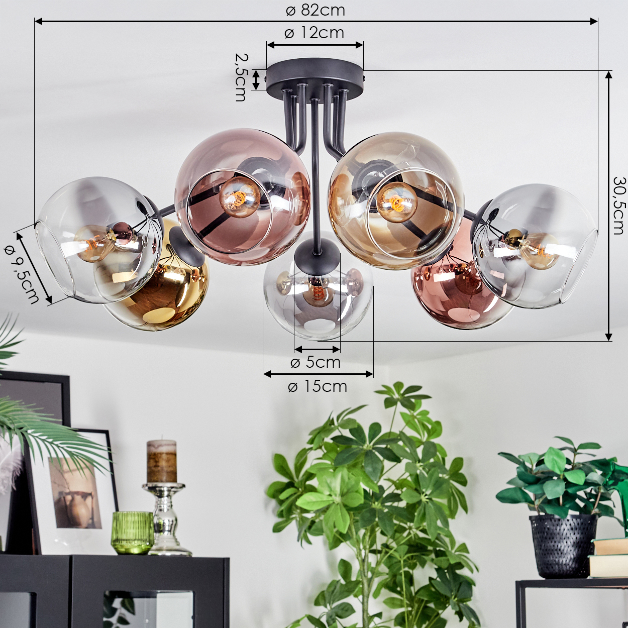 Eine moderne Deckenlampe mit sechs Glaskugeln in verschiedenen Farben. Schwarze Halterung. Grüne Pflanzen und ein schwarzer Schrank im Hintergrund.