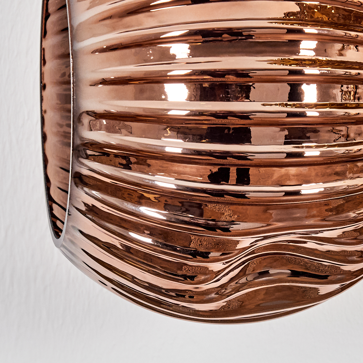 Nahaufnahme eines bronzefarbenen, wellenförmigen, reflektierenden Metallobjekts. Die Oberfläche reflektiert Licht und erzeugt Glanzlichter. Der Hintergrund ist eine weiße Wand.