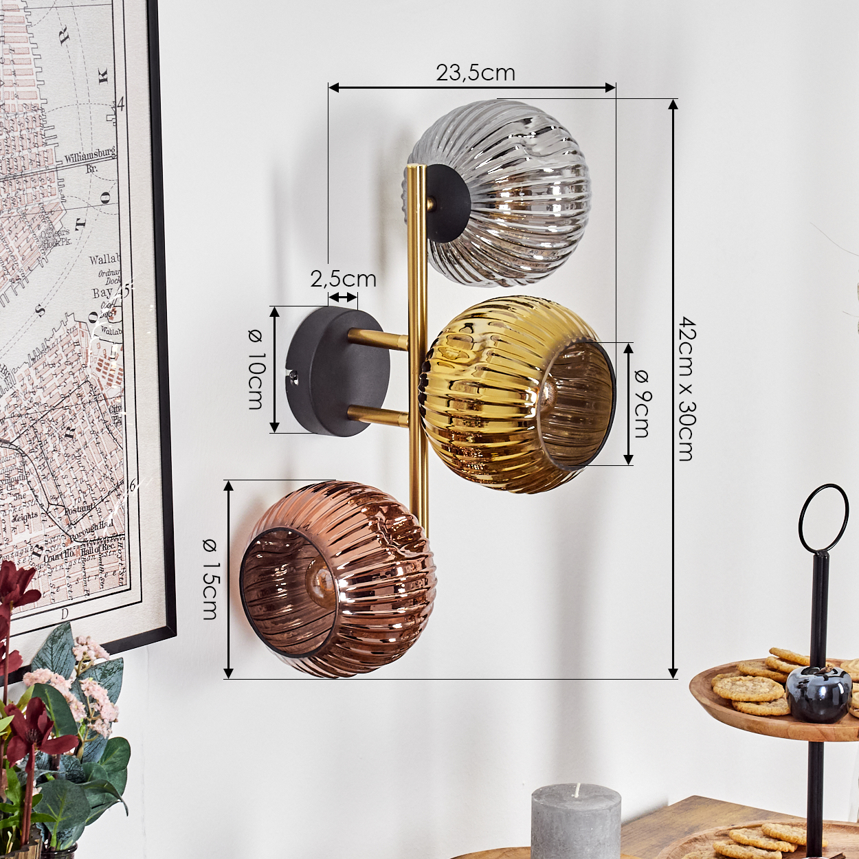 Eine Wandlampe mit drei Glaskugeln in Silber, Gold und Kupfer. Sie ist an einer weißen Wand mit einer gerahmten Karte links befestigt.