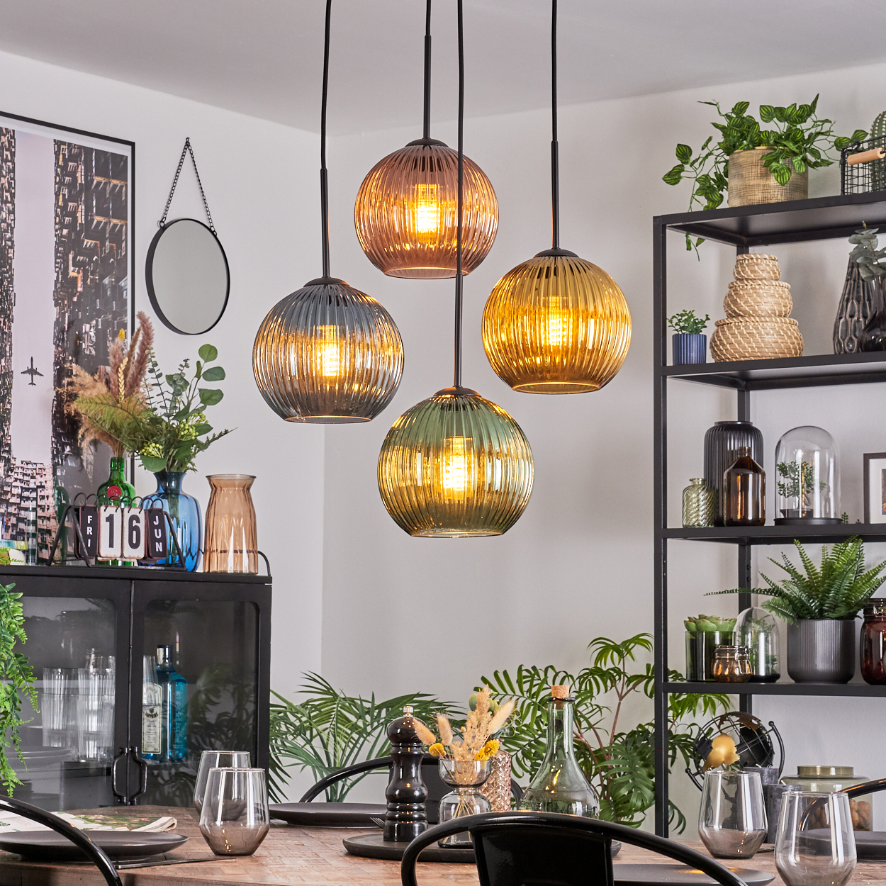 Esszimmer mit vier kugelförmigen Hängelampen. Pflanzen und Regale mit Dekoration vor weißer Wand. Tisch mit Gläsern gedeckt.