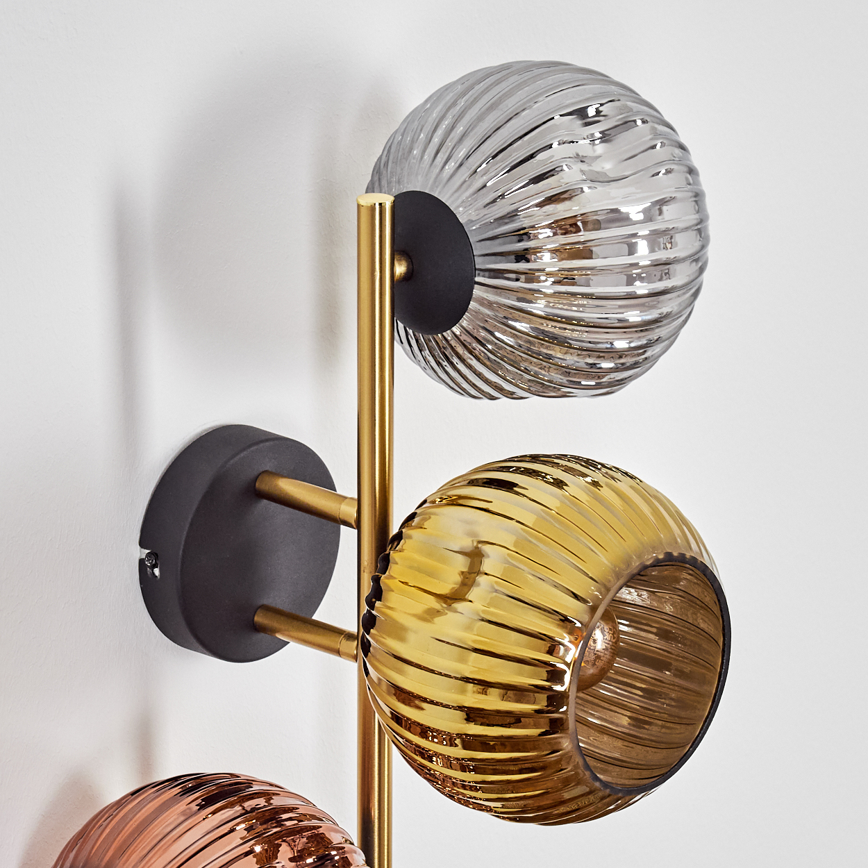 Eine moderne Wandleuchte mit drei Glaskugeln in Silber, Gold und Kupfer. Sie hat ein Messinggestell und einen schwarzen Sockel, montiert an einer weißen Wand.
