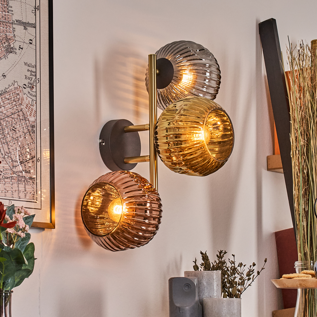 Eine Wandlampe mit drei Glaskugeln in Bernstein, Gold und Grau. Eine Karte und Pflanzen sind im Hintergrund sichtbar.