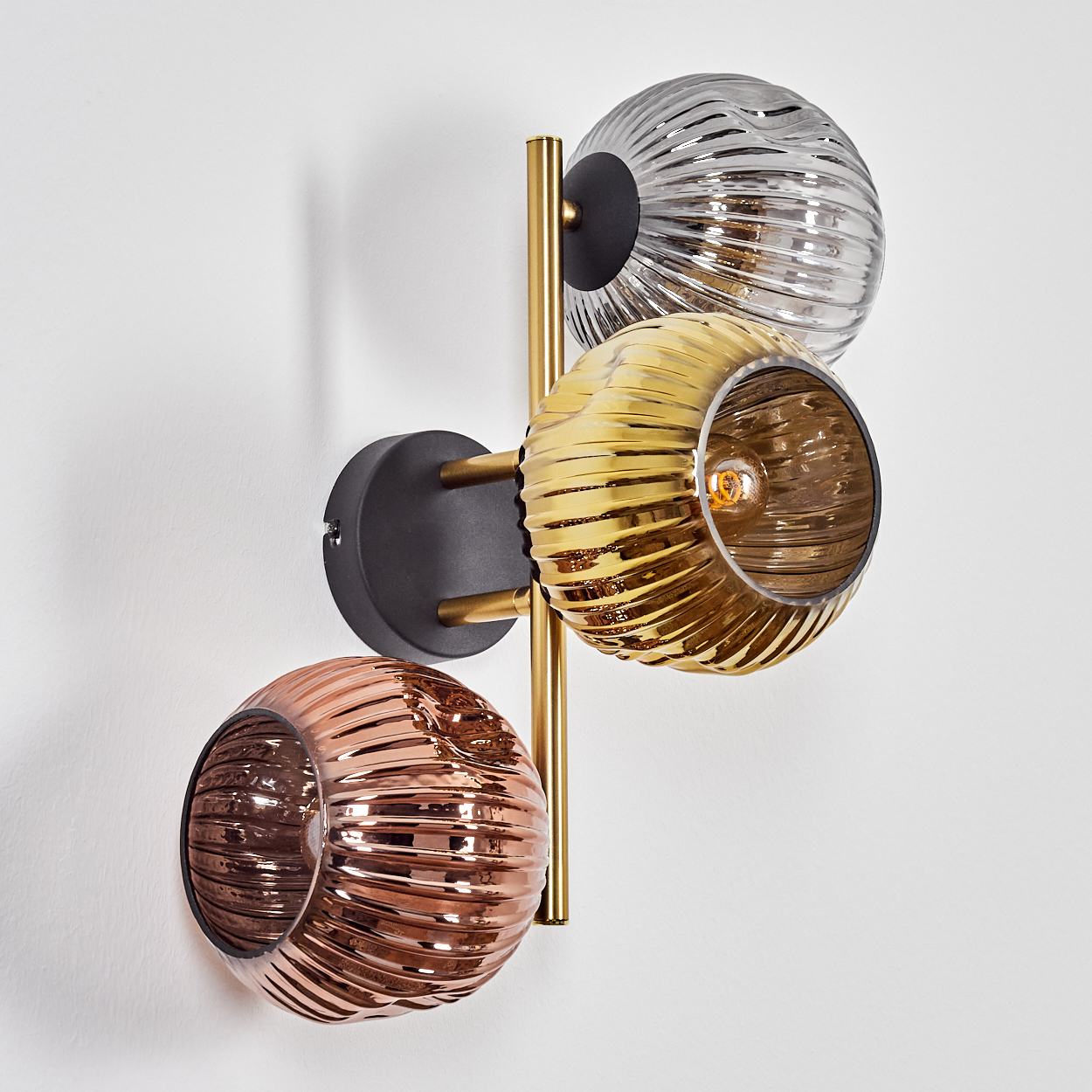 Eine Wandlampe mit drei Glaskugeln in Silber, Gold und Kupfer. Die Lampe ist an einer weißen Wand montiert. Die Kugeln haben ein Rippendesign.