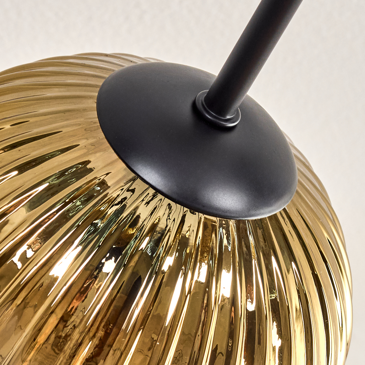 Nahaufnahme eines goldenen, gerippten Glasschirms mit schwarzem Metallaufsatz und Arm vor weißem Hintergrund.