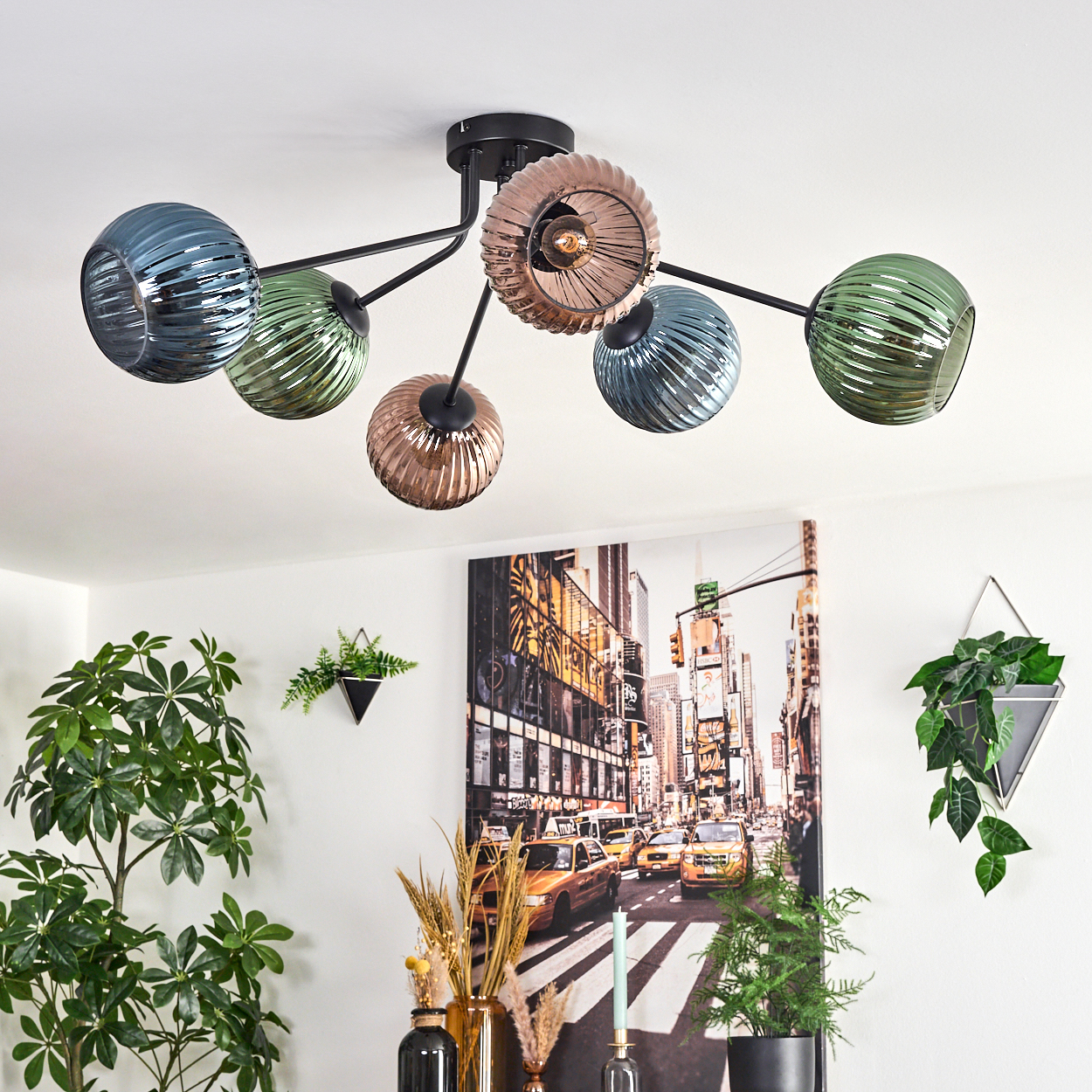 Ein moderner Kronleuchter mit bunten Glaskugeln hängt über einem Raum mit Pflanzen und einem Stadtbild-Kunstwerk.