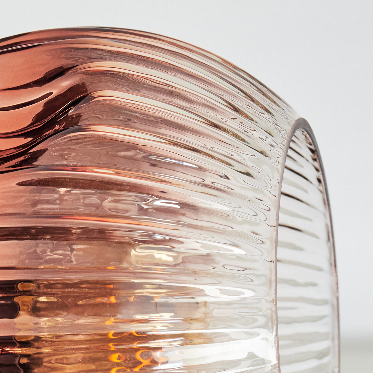 Nahaufnahme eines Glasobjekts mit welliger, gerippter Textur. Das Glas hat klare und rosafarbene Farbtöne mit Lichtreflexen.