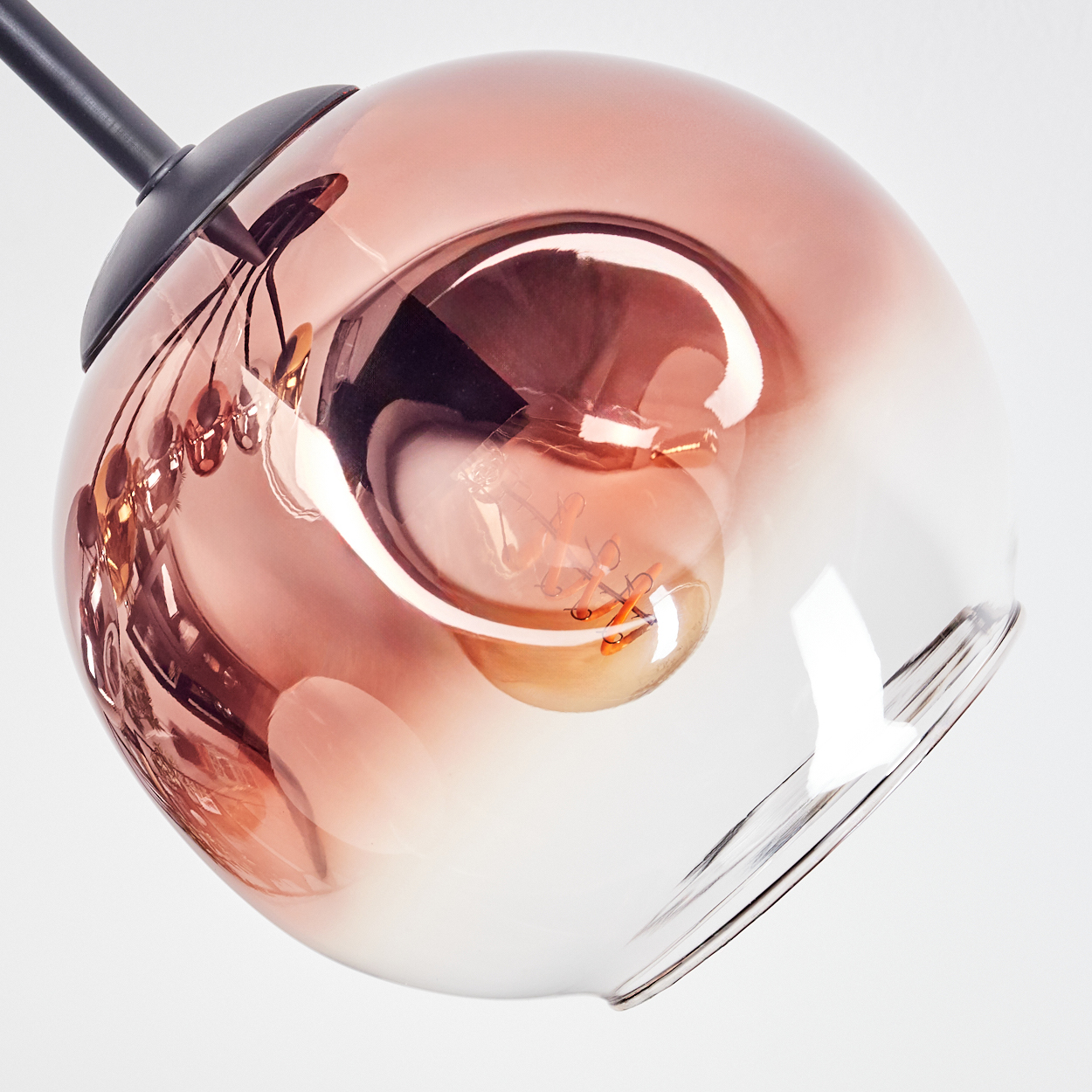 Nahaufnahme einer Glas-Pendelleuchte. Die Glühbirne im Inneren ist durch das roségoldfarbene Glas sichtbar. Ein schwarzer Stiel hält die Leuchte.