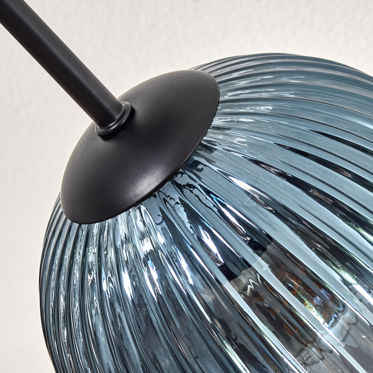 Nahaufnahme einer Lampe. Mit schwarzem Stiel und blauem, geripptem Glasschirm. Der Hintergrund ist eine schlichte, weiße Wand.