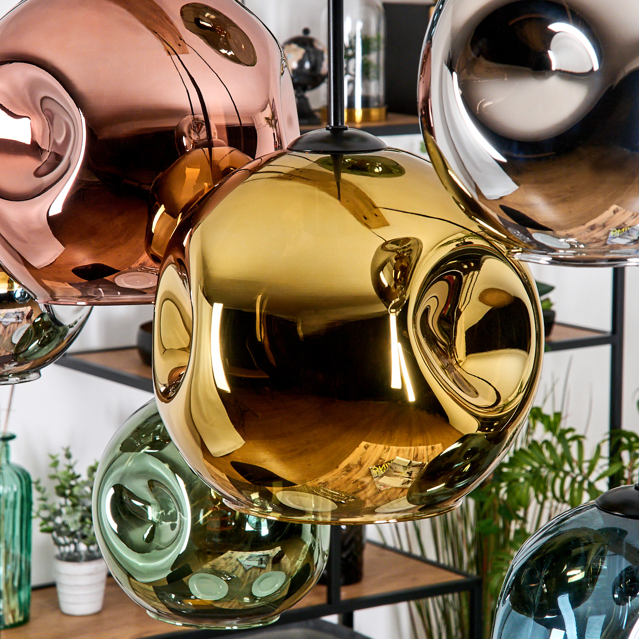 Nahaufnahme von modernen, reflektierenden, kugelförmigen Pendelleuchten in Kupfer, Gold, Grün und Silber. Im Hintergrund Regale mit Pflanzen.