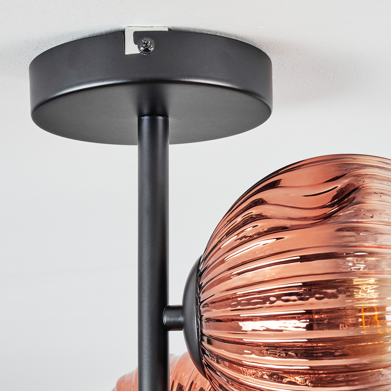 Nahaufnahme einer Deckenlampe. Sie hat einen schwarzen Metallrahmen und einen kupferfarbenen, strukturierten Glasschirm. Der Hintergrund ist weiß.