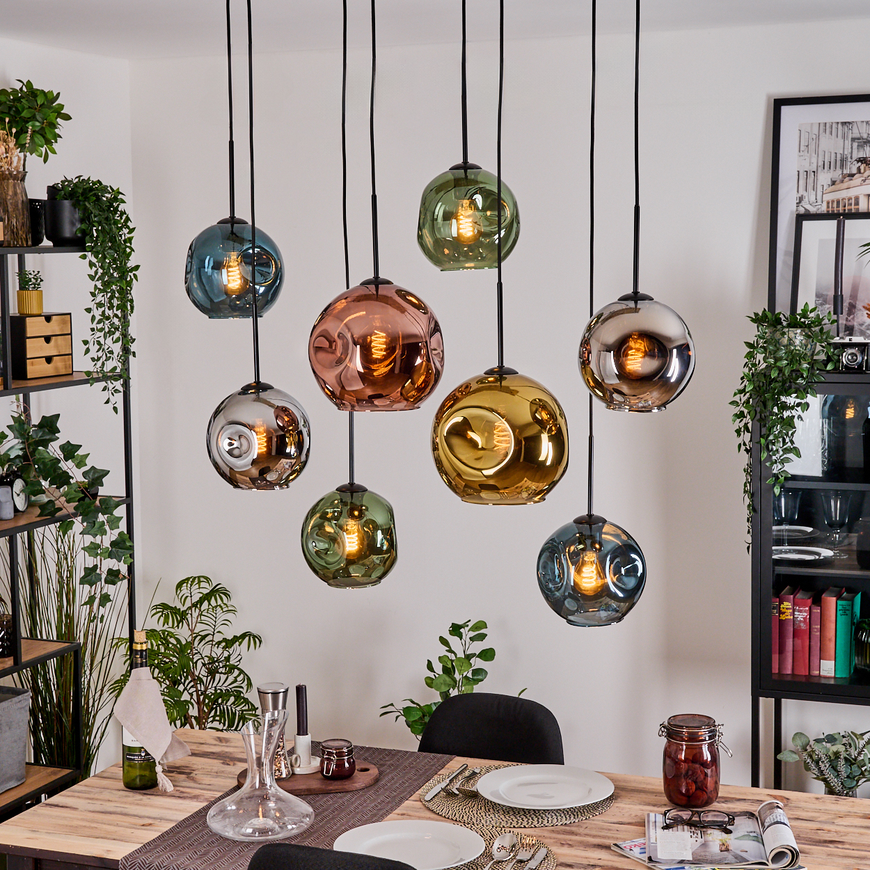 Esszimmer mit Holztisch, hängenden farbigen Kugelleuchten, Pflanzen und einem schwarz gerahmten Regal.