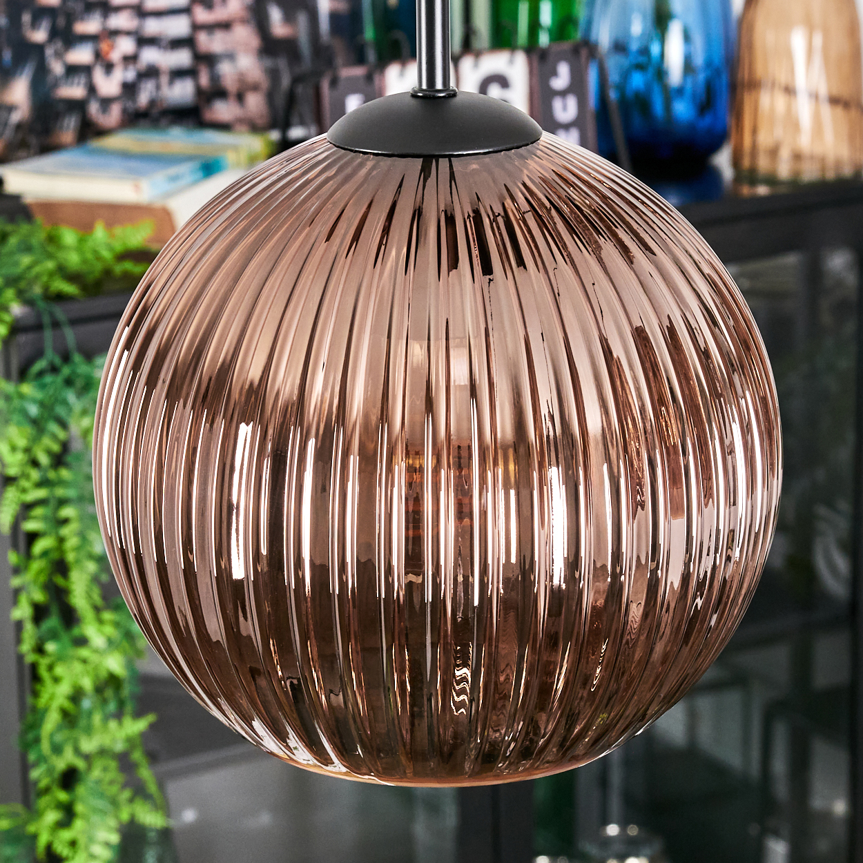Eine kupferfarbene, gerippte Glas-Pendelleuchte hängt und reflektiert Licht. Im Hintergrund sind grüne Pflanzen und verschwommene Objekte zu sehen.
