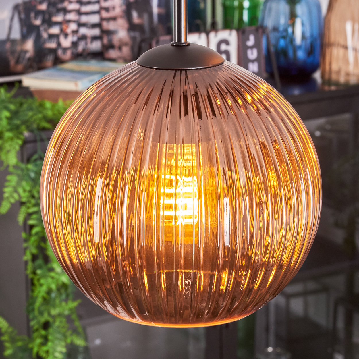 Nahaufnahme einer kupferfarbenen, gerippten Glas-Pendelleuchte mit einer leuchtenden Glühbirne. Hintergrund: grüne Pflanzen und Dekoration.