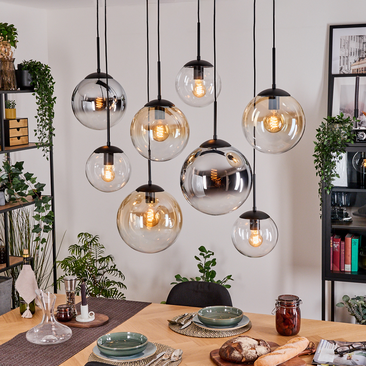 Esszimmer mit hängenden Kugelleuchten. Tisch mit Tellern, Brot und einem Glas. Pflanzen und Regale im Hintergrund.