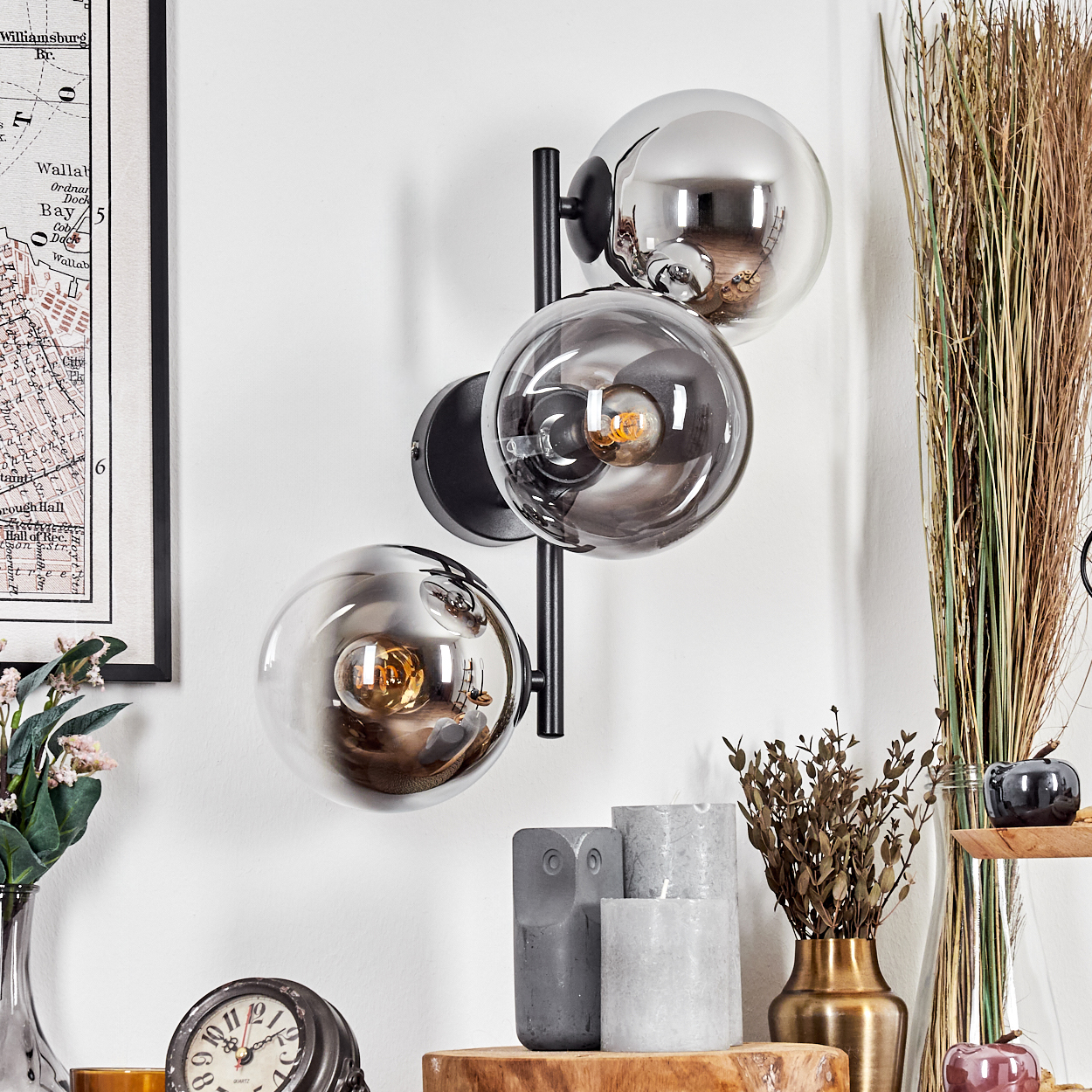Moderne Wandlampe mit drei Glaskugeln. Im Hintergrund eine Karte, Trockenpflanzen, Kerzen und eine Uhr.