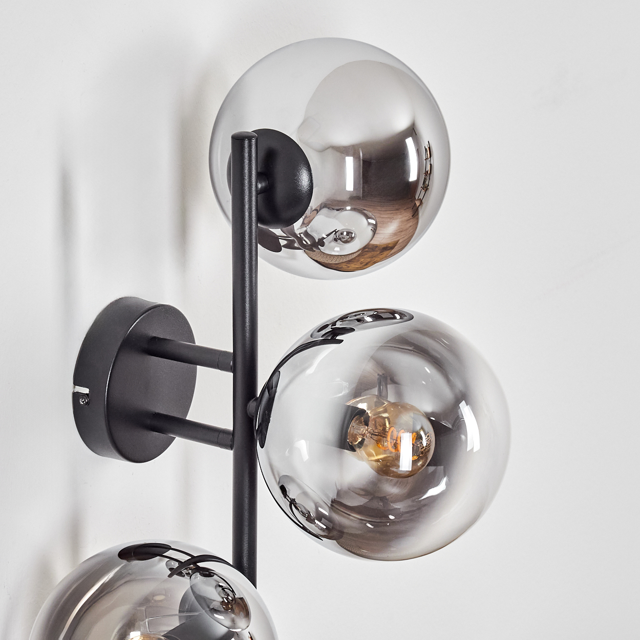 Eine moderne Wandleuchte mit drei kugelförmigen Glasschirmen. Die Lampe hat einen schwarzen Metallrahmen. Die Glühbirnen sind in den Glaskugeln sichtbar.