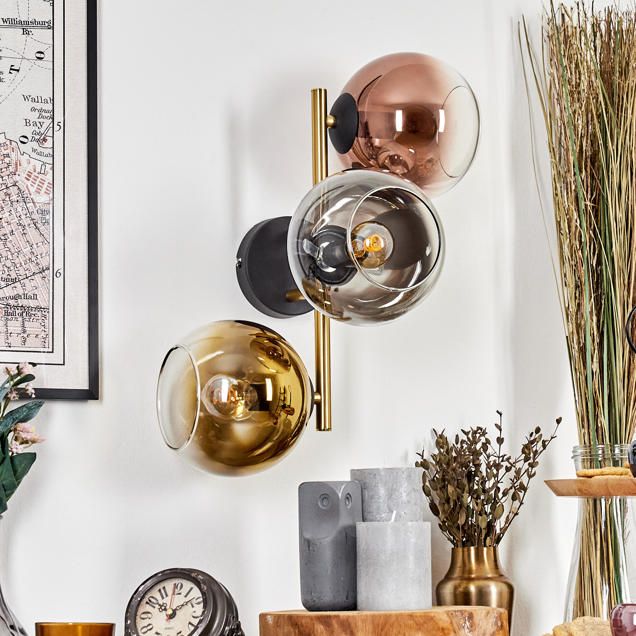 Eine moderne Wandlampe mit drei Glaskugeln. Im Hintergrund eine Karte, Trockenpflanzen, Kerzen und eine Uhr.