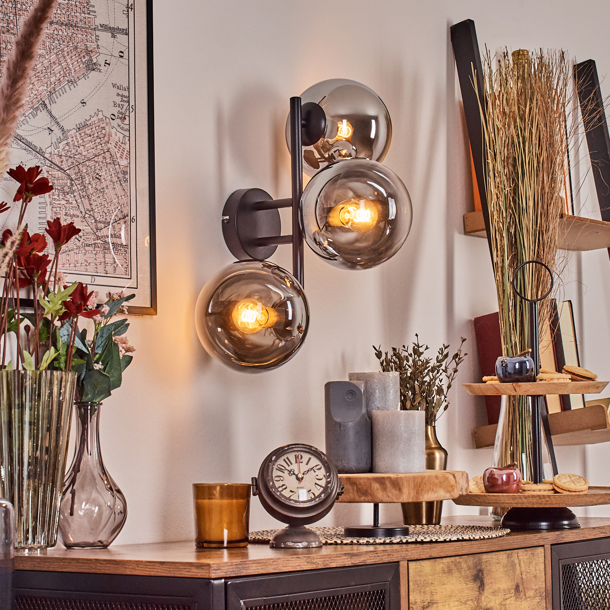 Ein Raum mit Wandlampe, Uhr und Tisch mit Dekorationen. Eine Karte an der Wand. Blumen und Trockenpflanzen vorhanden.