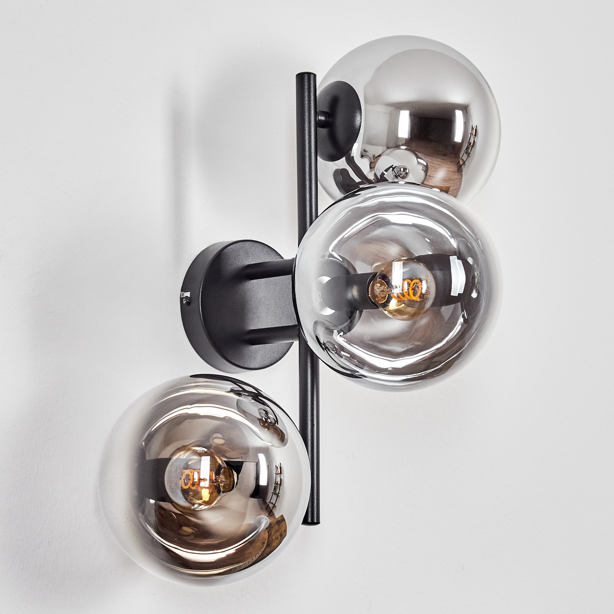 Eine moderne Wandlampe mit drei silbernen Glaskugeln und schwarzen Metallarmen. Glühbirnen sind sichtbar. Die Wand ist weiß.