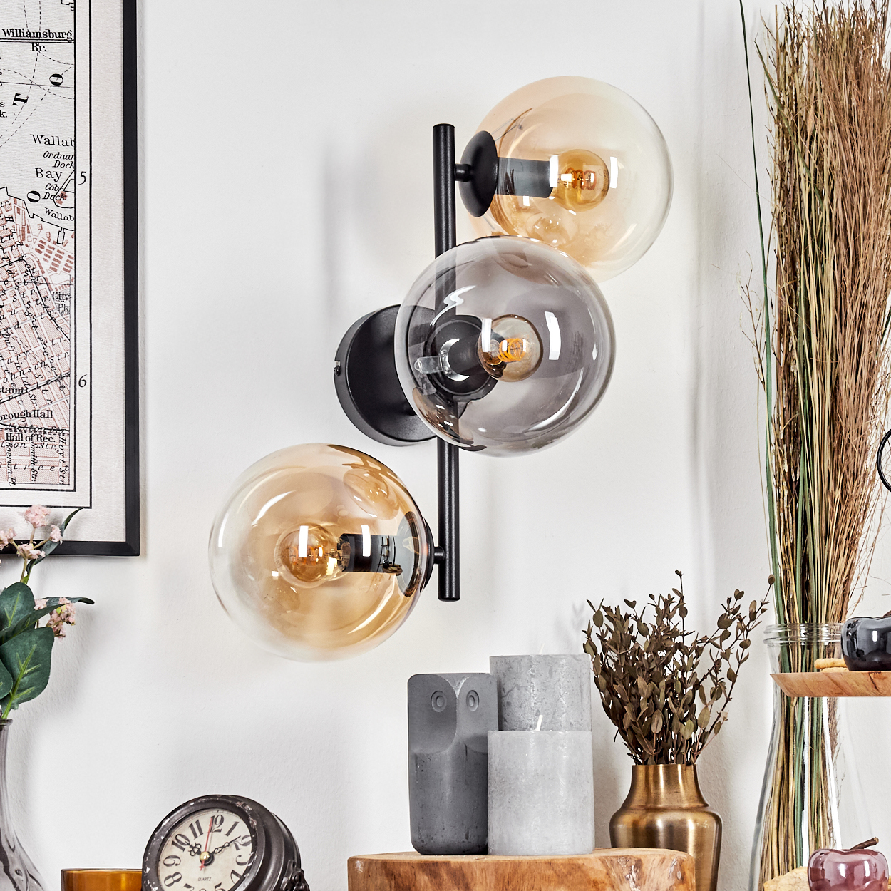 Eine moderne Wandlampe mit drei Glaskugeln. Verschiedene Dekorationen, darunter Kerzen, Uhr und Trockenpflanzen, sind sichtbar.