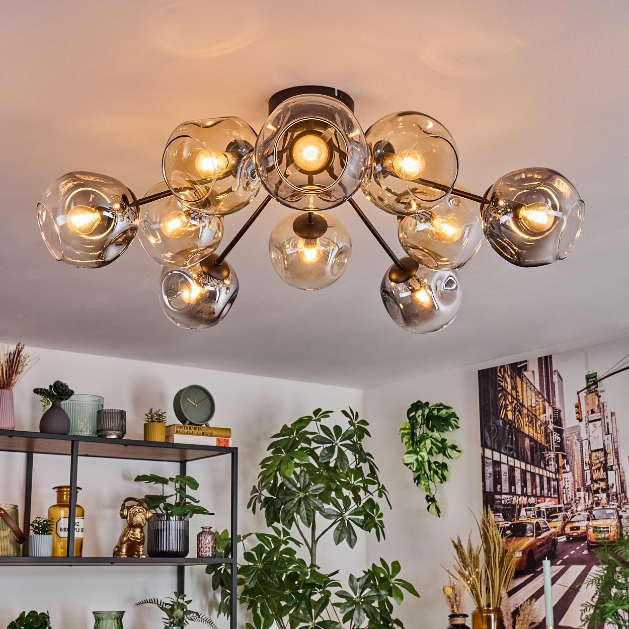 Eine moderne Deckenleuchte mit Glaskugeln hängt. Darunter ein Regal mit Dekoration und Pflanzen. Ein Gemälde von New York City an der Wand.