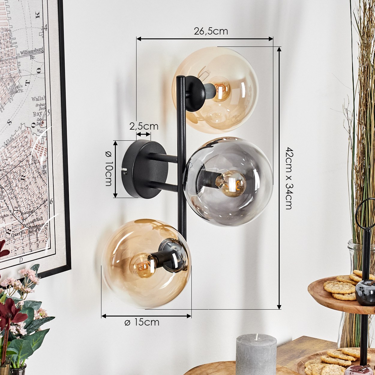 Eine moderne Wandlampe mit drei kugelförmigen Schirmen. Maße sind markiert. Im Hintergrund sind eine Karte, Blumen und ein Serviertablett.