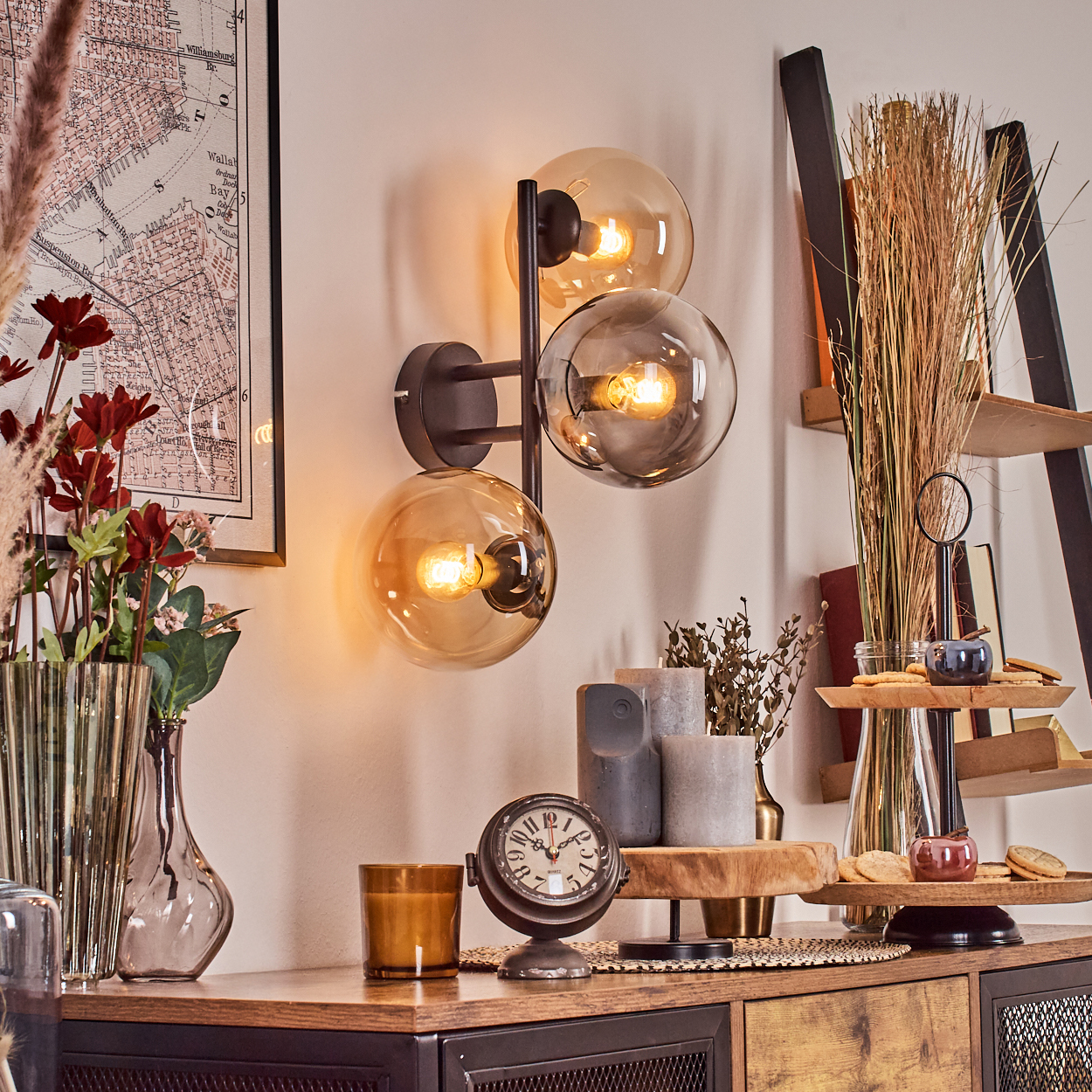 Dekorative Wandlampe mit drei Glaskugeln. Ein Holzschrank mit Dekorartikeln wie einer Uhr, Kerzen und einem Tablett mit Keksen.