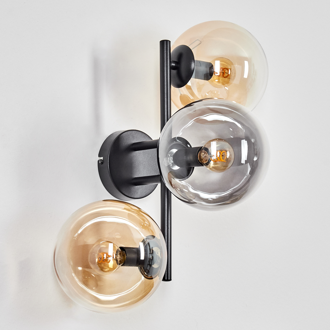 Moderne Wandlampe mit drei Glaskugeln. Zwei Kugeln sind bernsteinfarben, eine ist klar. Schwarzer Metallrahmen. An einer weißen Wand montiert.