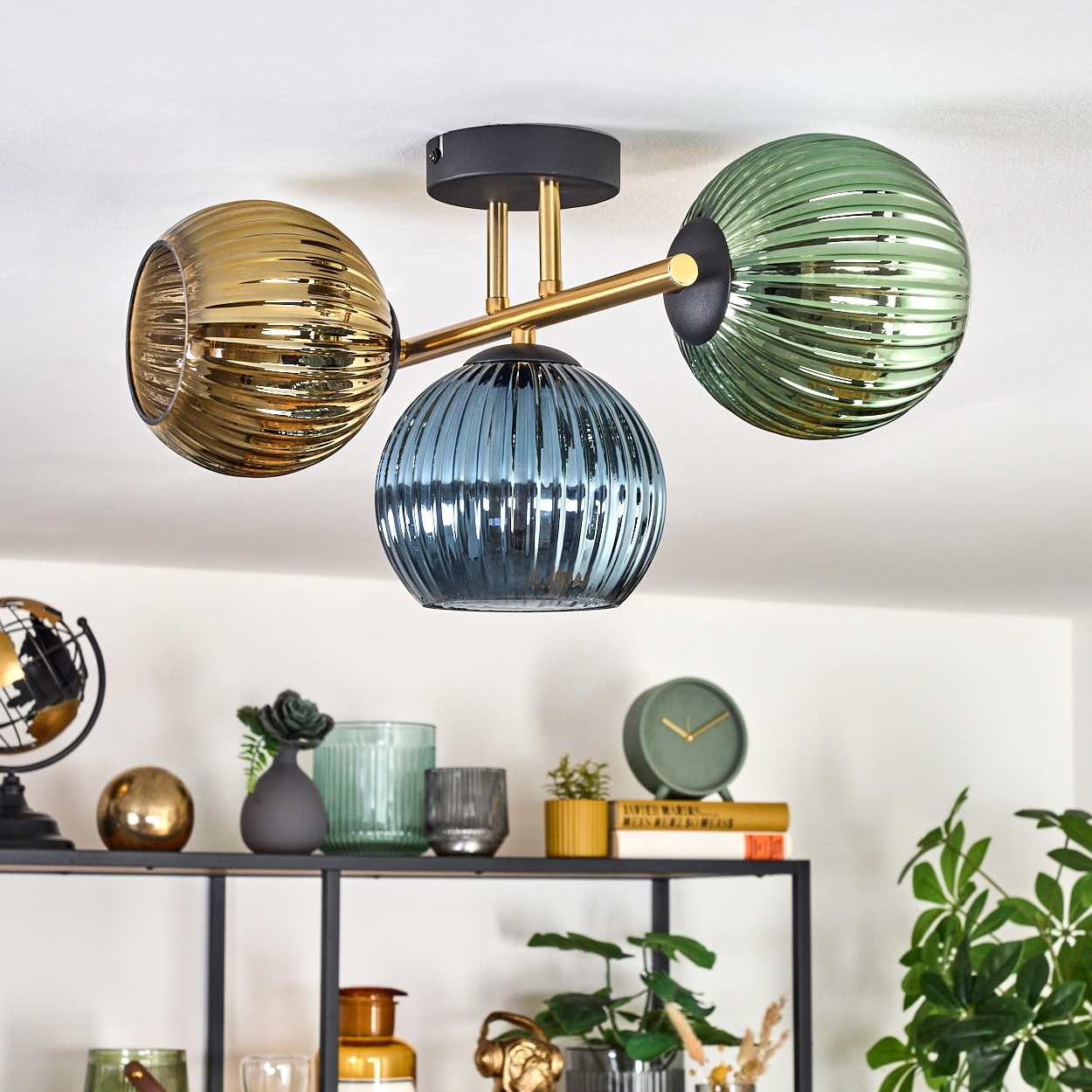 Eine Deckenleuchte mit drei Glaskugeln in Gold, Blau und Grün, über einem Regal mit Dekoration, darunter ein Globus und eine Uhr.