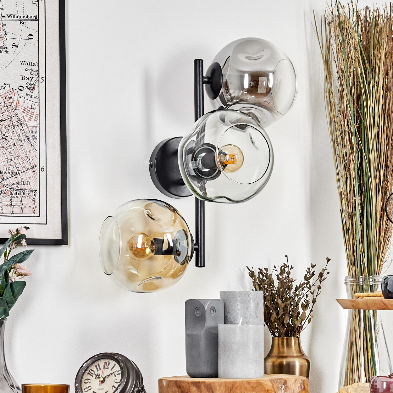 Eine Wandlampe mit drei Glaskugeln, einer Karte, Kerzen und Dekorationsartikeln. Die Lampe hat einen schwarzen Rahmen. Der Hintergrund ist weiß.