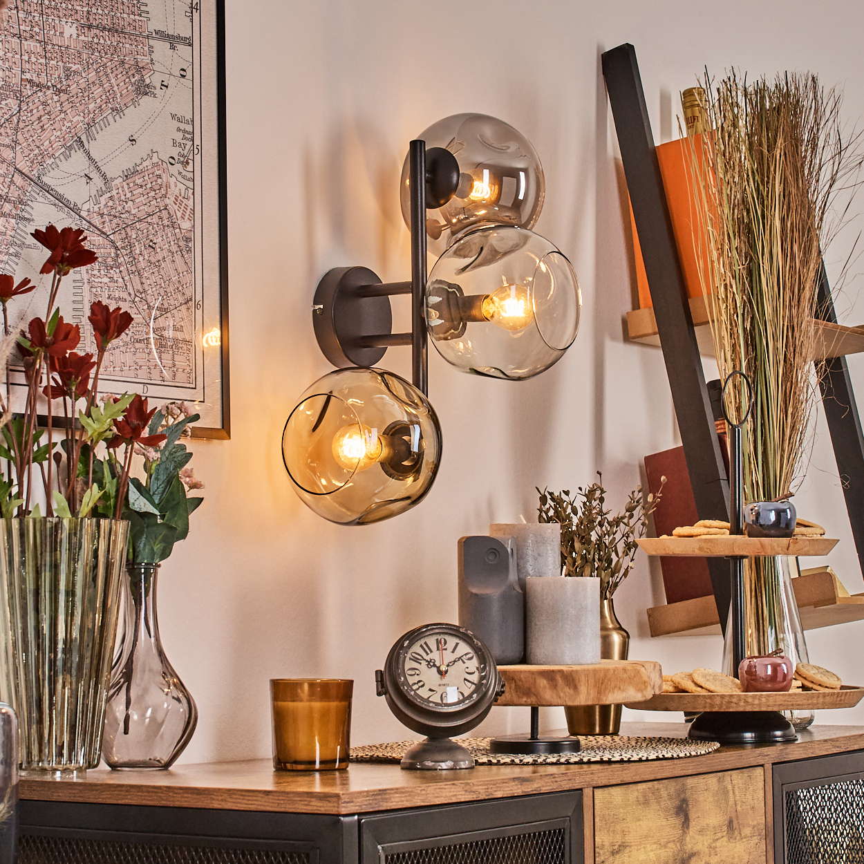 Eine dekorative Szene mit Wandlampe, Uhr, Kerzen, Blumen und einer Karte. Holzmöbel und ein Leiterregal sind ebenfalls vorhanden.