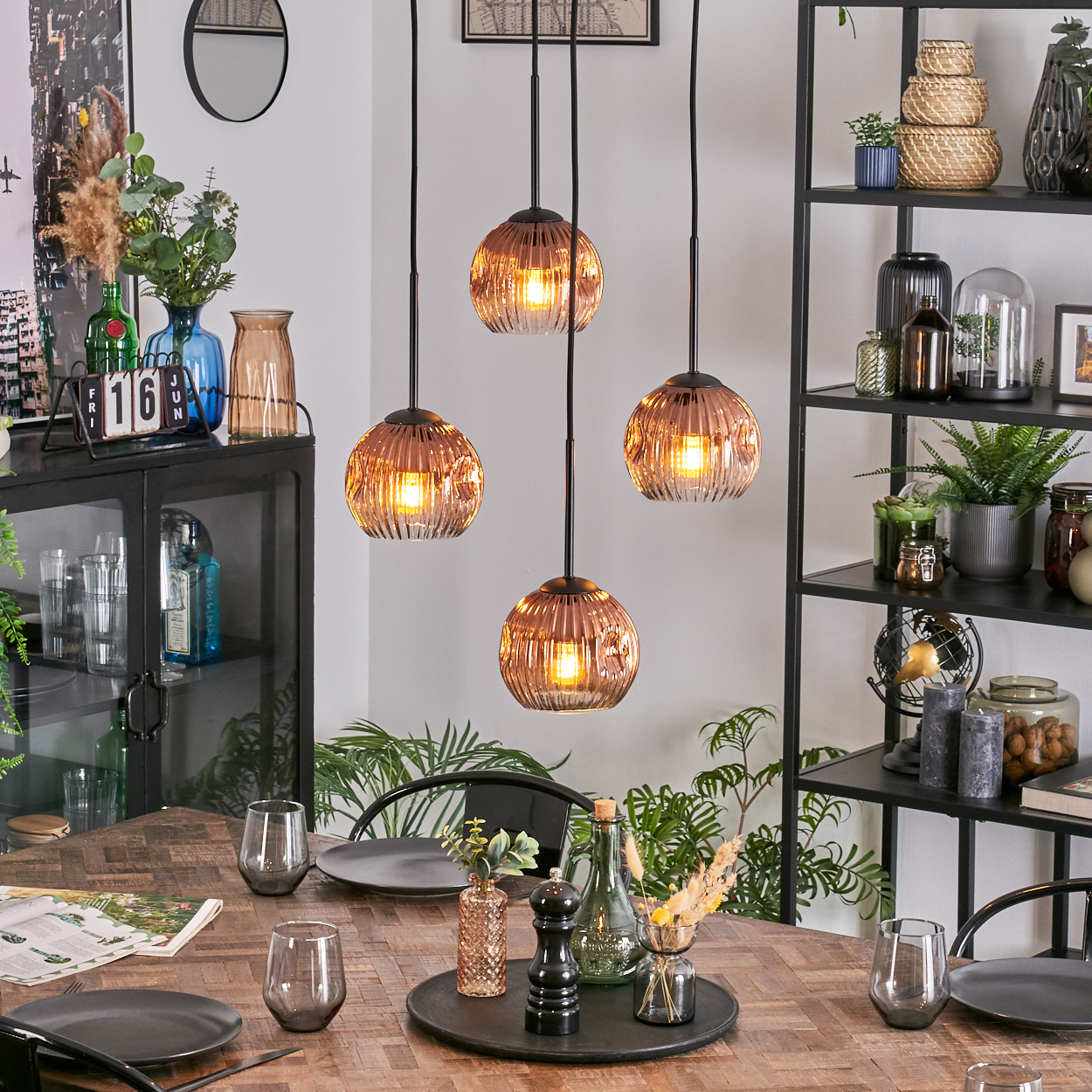 Esszimmer mit Tisch für vier Personen. Hängelampen, ein schwarzes Regal mit Dekoration und ein Schrank mit Glastüren sind zu sehen.