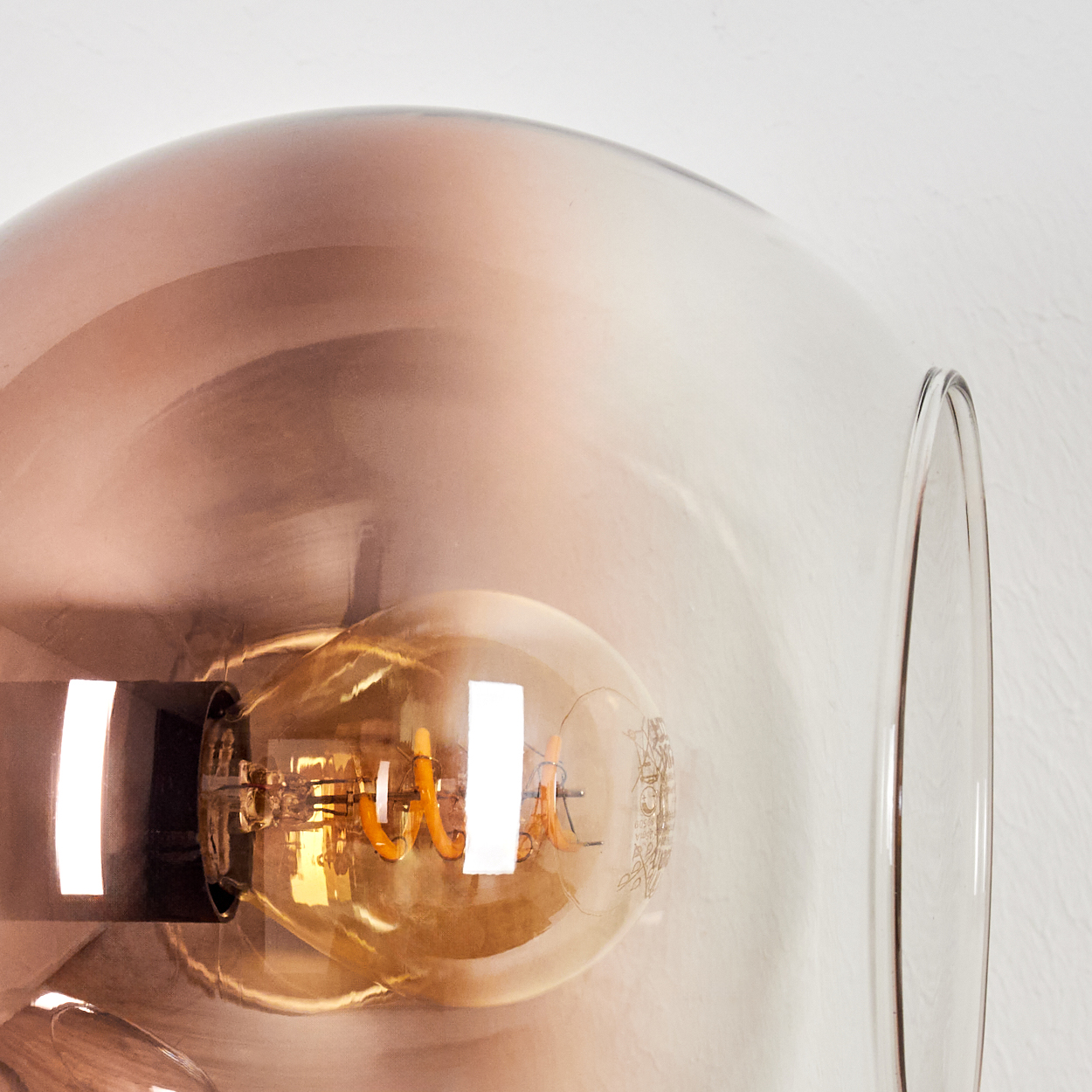 Nahaufnahme einer Leuchte mit Glasschirm. Im Inneren ist eine Glühbirne mit einem gewendelten Glühfaden zu sehen. Der Hintergrund ist eine weiße Wand.