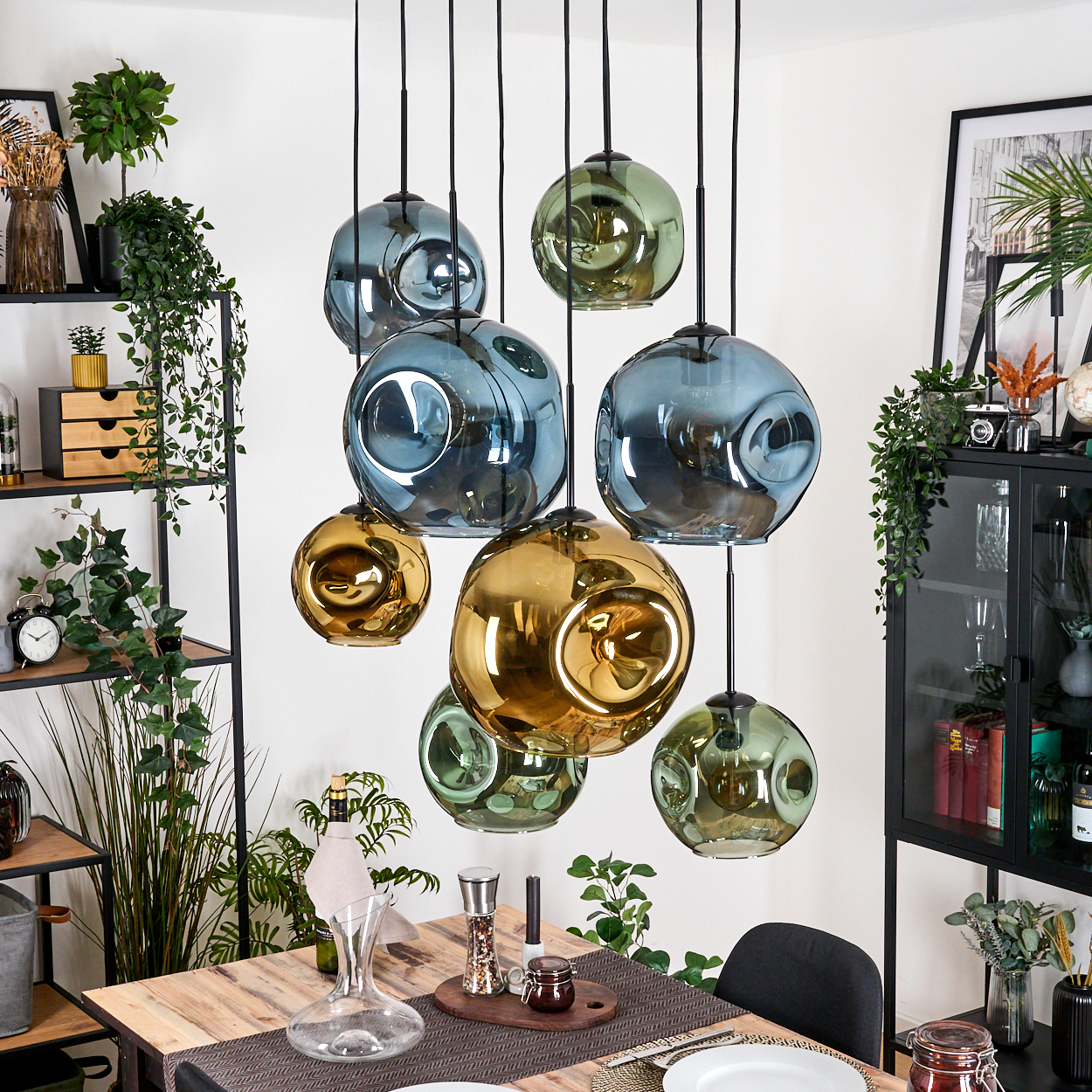 Ein Esszimmer mit hängenden Glaskugelleuchten in Gold, Blau und Grün. Ein Holztisch ist mit Dekoration und einer Weindekantierkanne gedeckt.
