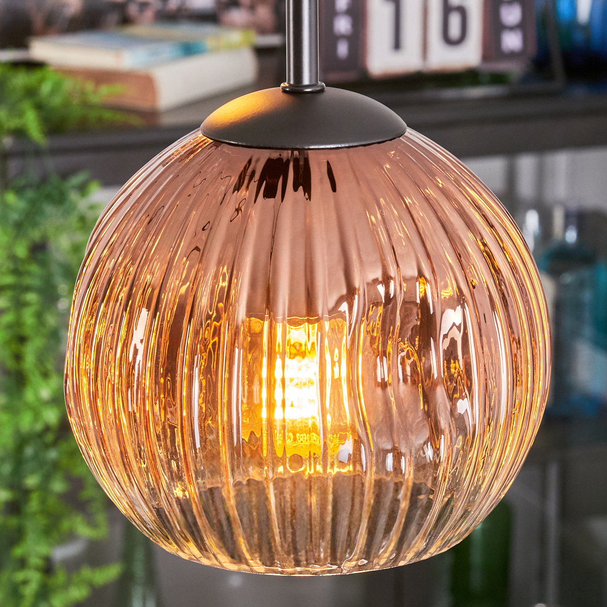 Nahaufnahme einer Glas-Pendelleuchte mit warmem Schein. Das Licht hat einen kupferfarbenen Farbton und eine geriffelte Textur. Der Hintergrund umfasst Pflanzen.