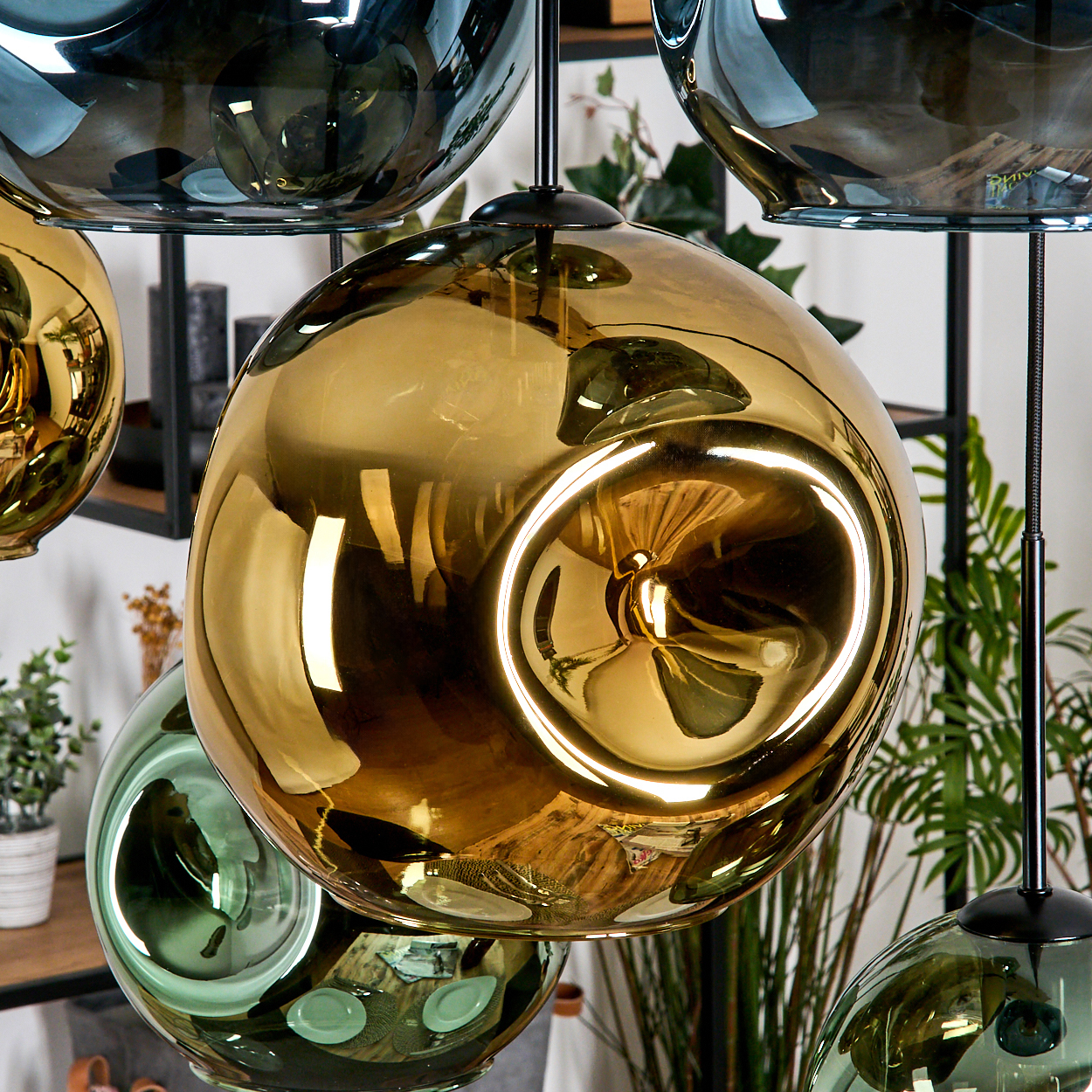 Nahaufnahme von goldenen und tealfarbenen Glas-Pendelleuchten. Die Lichter reflektieren den Raum. Pflanzen und Dekoration sind sichtbar.