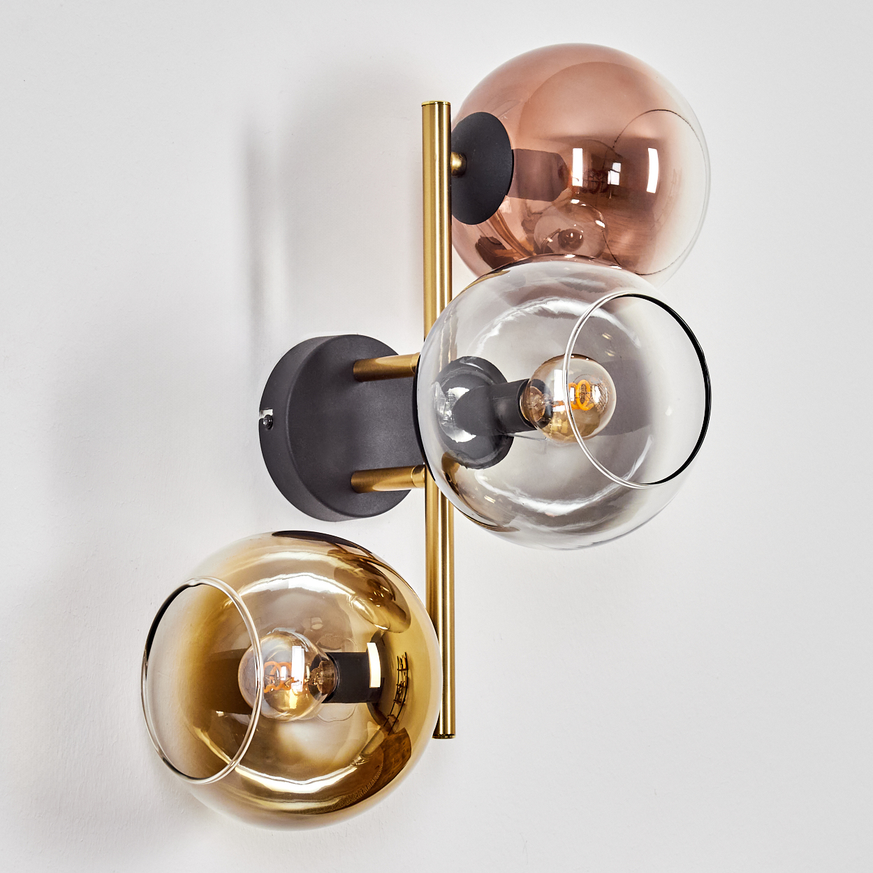 Eine moderne Wandlampe mit drei Glaskugelschirmen in Kupfer, Klar und Gold. Sie hat einen schwarzen Sockel und goldene Akzente, an einer weißen Wand montiert.