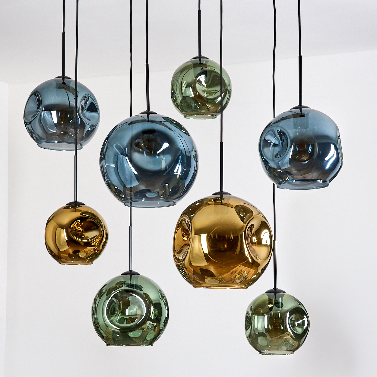 Neun kugelförmige Pendelleuchten hängen von der Decke. Sie sind in verschiedenen Farben gehalten, darunter Gold, Blau und Grün, vor einer weißen Wand.