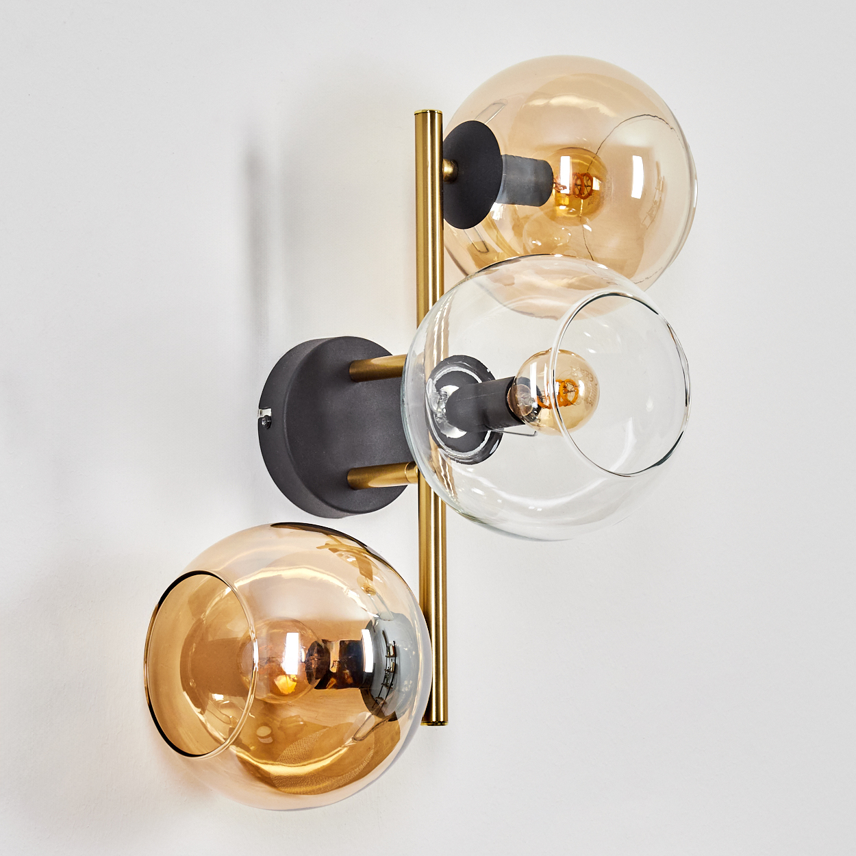 Eine moderne Wandlampe mit drei Glaskugeln. Zwei sind bernsteinfarben, eine klar. Schwarze und goldene Akzente an einer weißen Wand. Glühbirnen leuchten.
