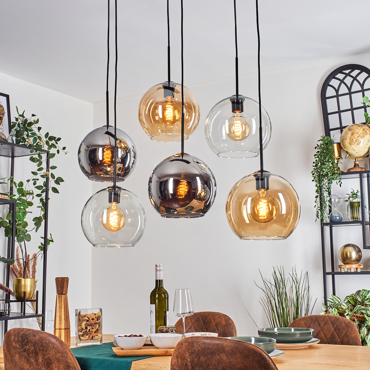 Esszimmer mit sechs hängenden Kugelleuchten, einem Holztisch mit Tellern, Wein und Stühlen. Regale und Pflanzen im Hintergrund.