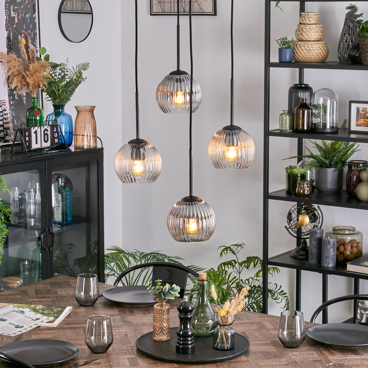 Esszimmer mit Holztisch, vier Hängelampen, Gläsern, Tellern und dekorativen Gegenständen auf Regalen und dem Tisch.