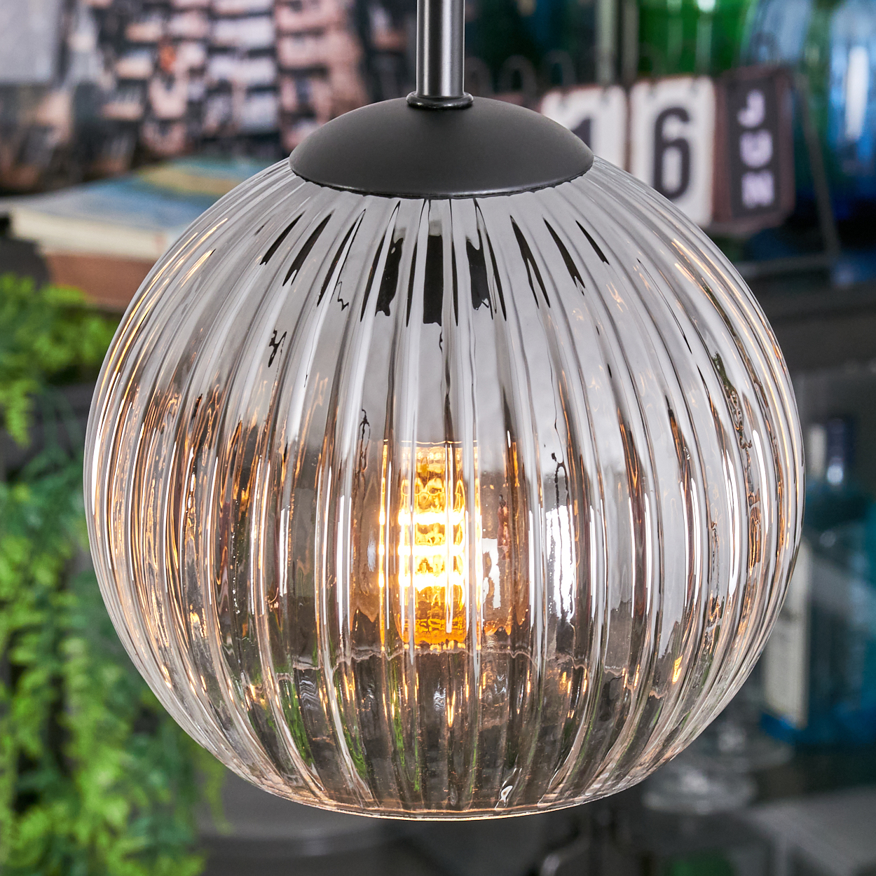 Eine klare, gerippte Glas-Pendelleuchte mit einer beleuchteten, warm getönten Glühbirne im Inneren. Der Hintergrund ist verschwommen.