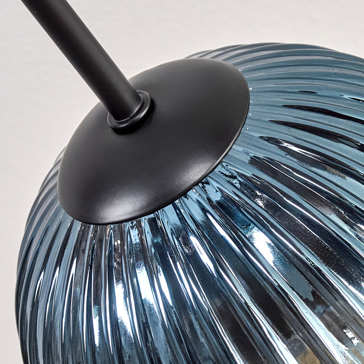 Nahaufnahme eines blauen Glaslampenschirms mit vertikalen Rippen, unter einer schwarzen Metallscheibe und -stange. Der Hintergrund ist weiß.