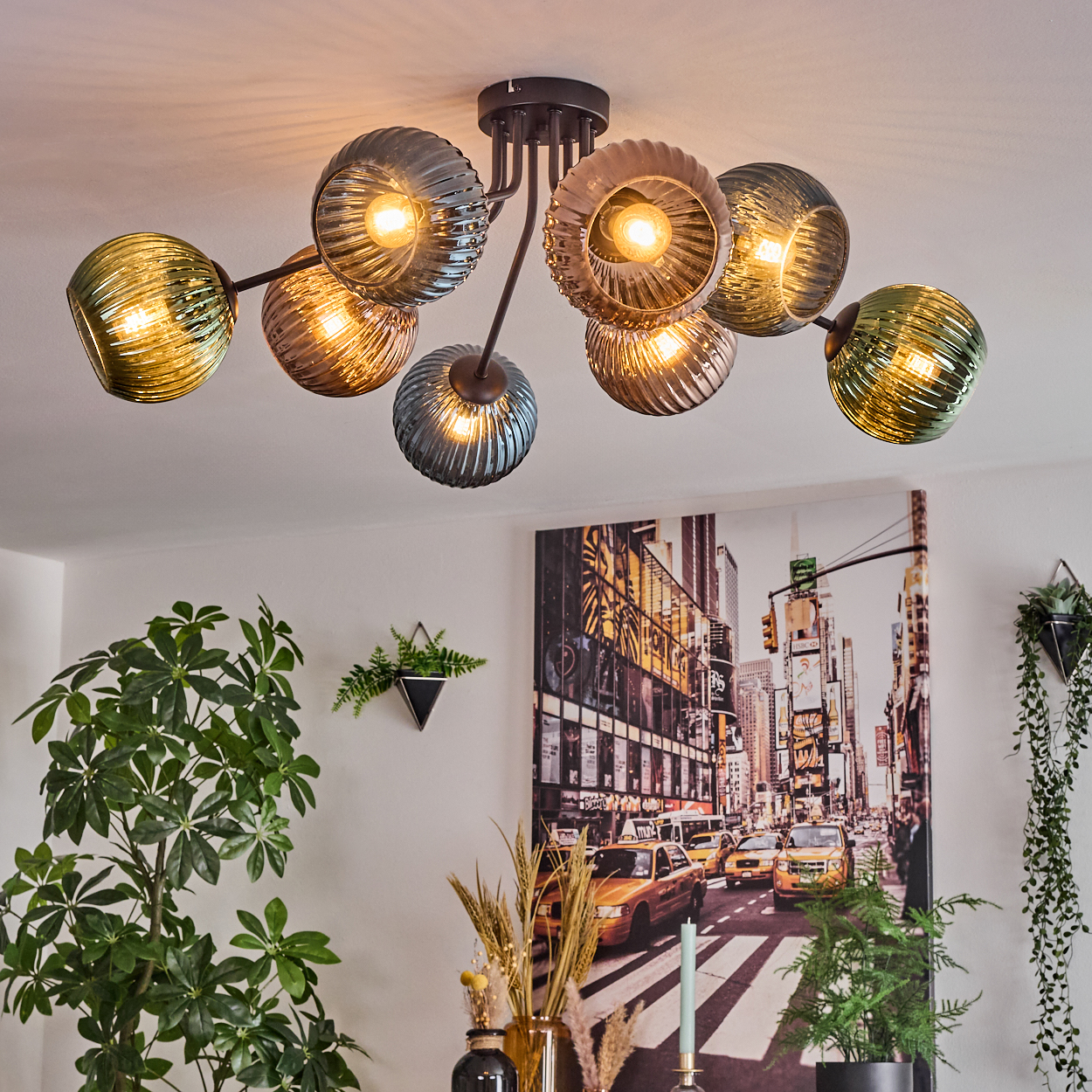 Eine moderne Deckenleuchte mit mehreren Glaskugeln beleuchtet einen Raum. Ein Stadtbildgemälde hängt an der Wand, Pflanzen im Vordergrund.