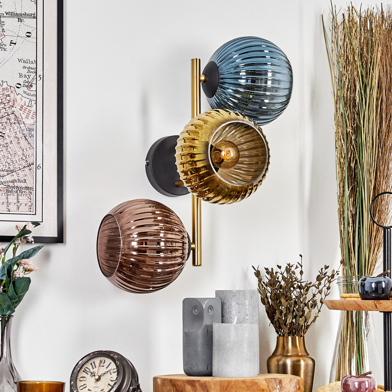 Eine Wandlampe mit drei Glaskugeln in Blau, Gold und Bronze ist an einer weißen Wand befestigt. Weitere Dekorationen umfassen eine Karte und Kerzen.