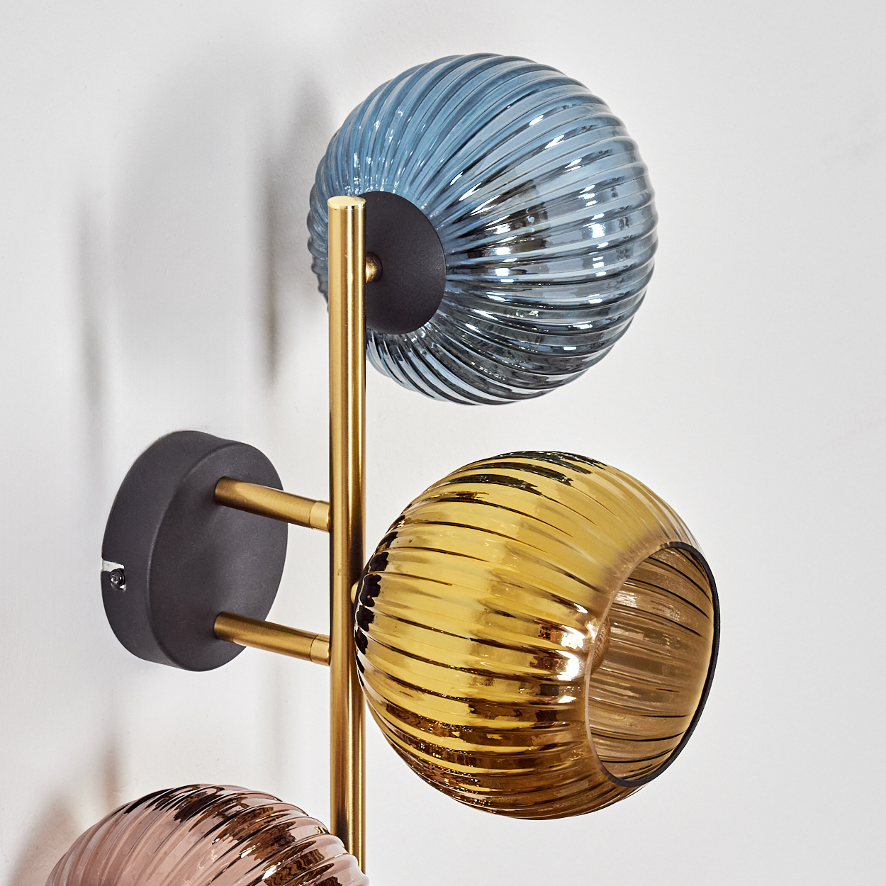 Eine moderne Wandleuchte mit drei kugelförmigen Lampenschirmen in Blau, Gold und Rosa, getragen von einem goldenen Rahmen, vor einer weißen Wand.