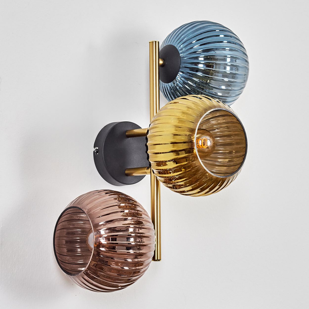 Eine Wandlampe mit drei Glaskugeln in Blau, Gelb und Braun, vertikal angeordnet. Die Halterung ist gold- und schwarzfarben, an einer weißen Wand.