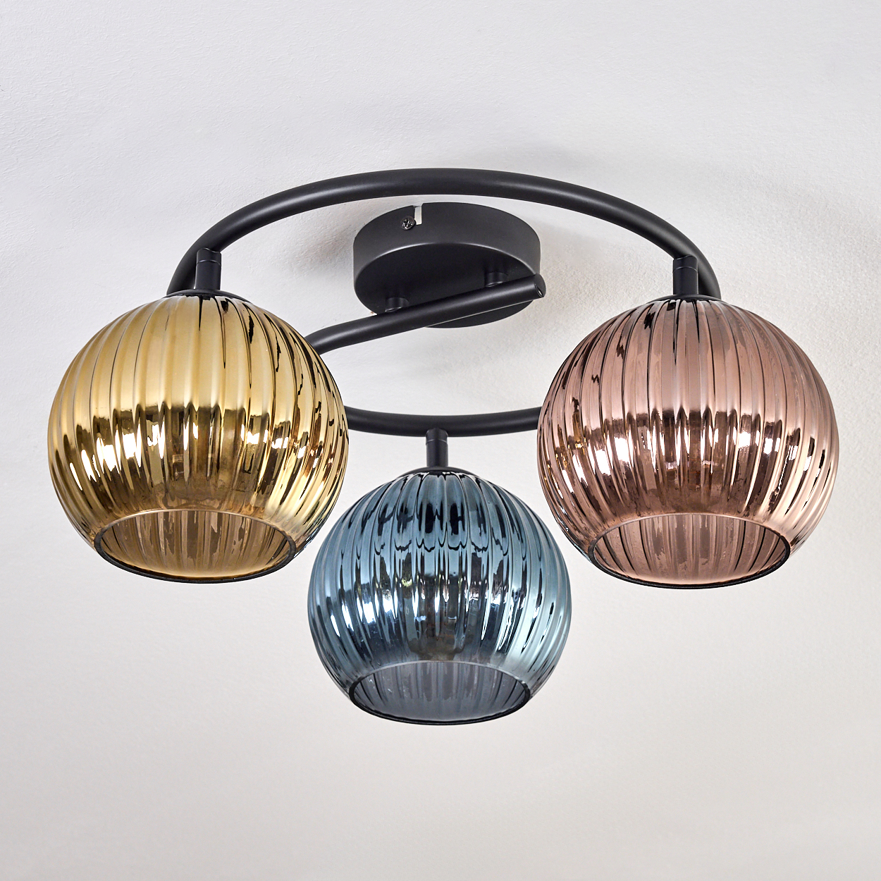 Eine moderne Deckenleuchte mit drei Glaskugeln in Gold, Blau und Kupfer. Die Leuchte ist schwarz und an einer weißen Decke montiert.